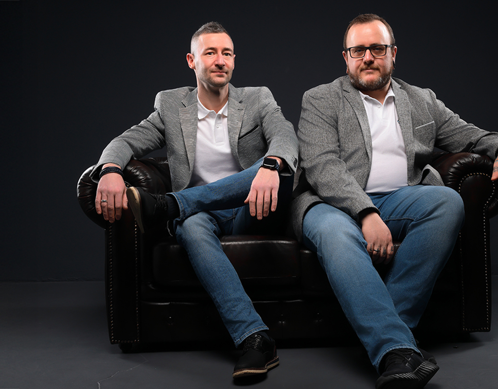 Daniel und Sascha Klein sitzen auf einem Ledersofa. Sie tragen beide graue Sakkos und weiße Poloshirts und blicken in die Kamera.